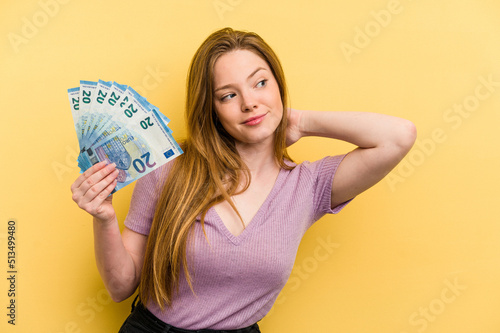 Young caucasian woman holding banknotes isolated on yellow background touching back of head, thinking and making a choice.