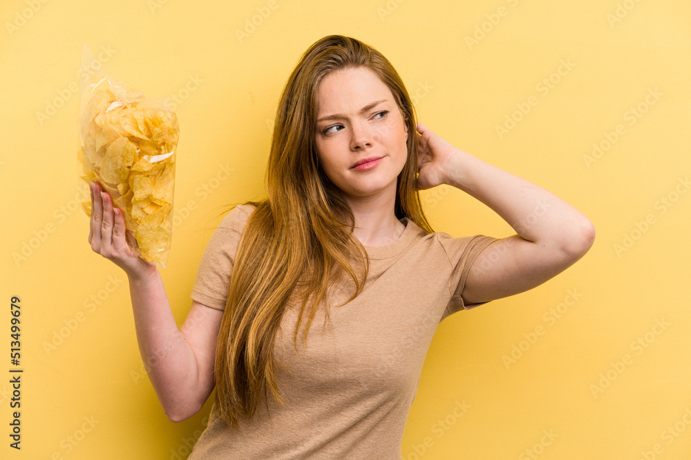 Young caucasian woman holding a bag of chips isolated on yellow background touching back of head, thinking and making a choice.
