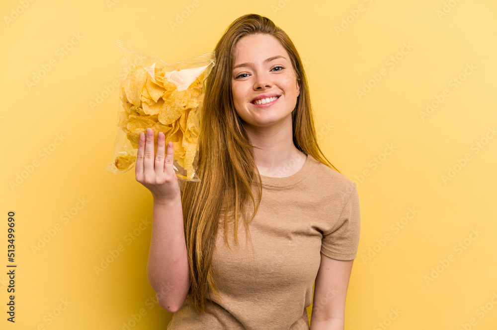 Young caucasian woman holding a bag of chips isolated on yellow background happy, smiling and cheerful. Young caucasian woman holding a bag of chips isolated on yellow background happy, smiling and cheerful.