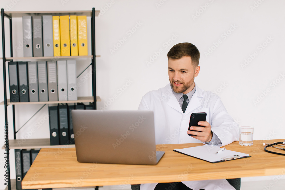 Male doctor holds a phone and sits in the office near the laptop Male doctor holds a phone and sits in the office near the laptop