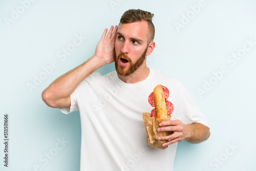 Young caucasian man eating a sandwich isolated on blue background trying to listening a gossip.