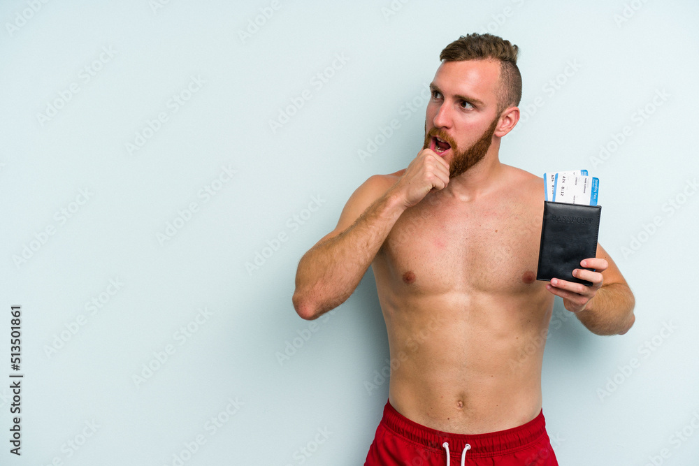Young caucasian man holding a passport isolated on blue background looking sideways with doubtful and skeptical expression. Young caucasian man holding a passport isolated on blue background looking sideways with doubtful and skeptical expression.