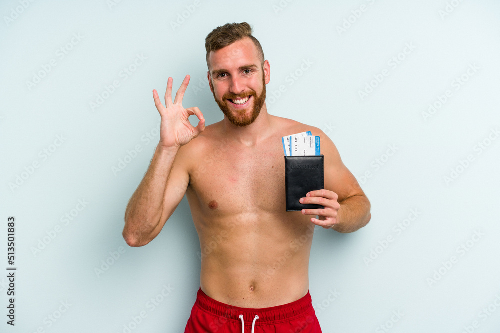 Young caucasian man holding a passport isolated on blue background cheerful and confident showing ok gesture. Young caucasian man holding a passport isolated on blue background cheerful and confident showing ok gesture.