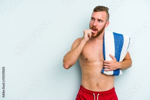 Young caucasian man going to the beach holding a towel isolated on blue background relaxed thinking about something looking at a copy space.