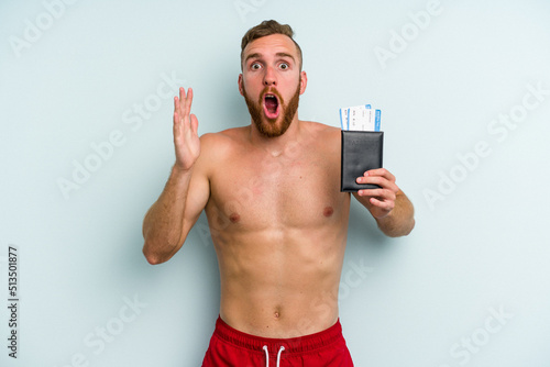 Young caucasian man holding a passport isolated on blue background surprised and shocked.