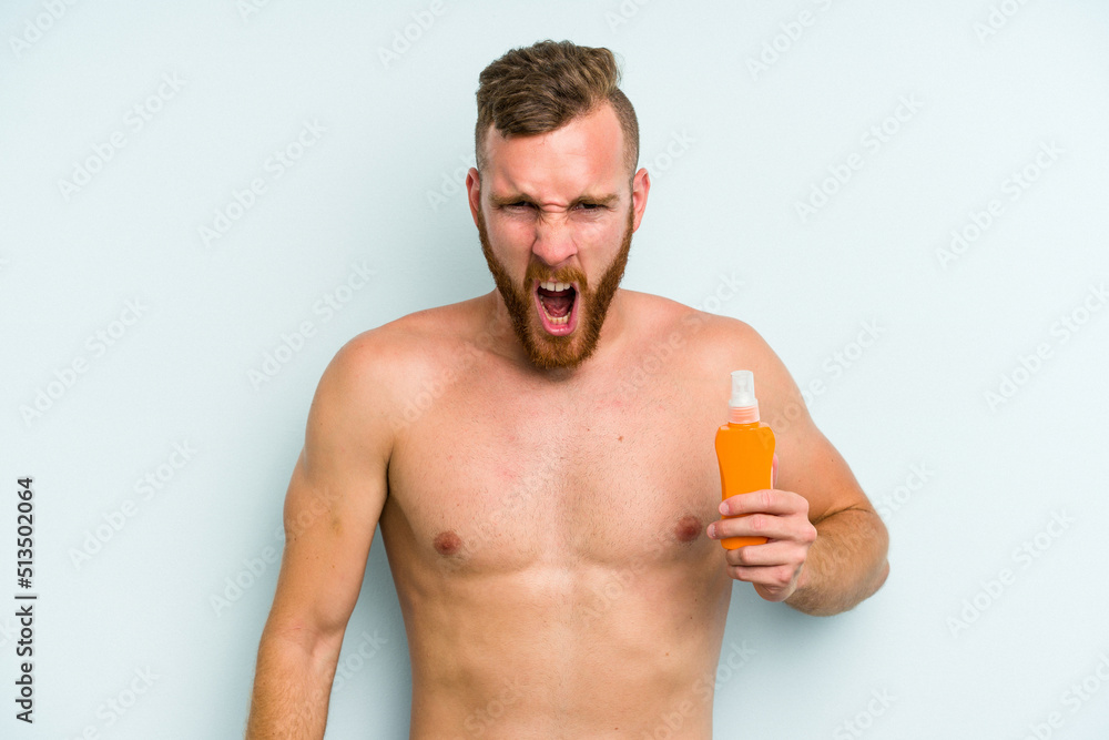 Young caucasian man holding solar cream isolated on blue background screaming very angry and aggressive. Young caucasian man holding solar cream isolated on blue background screaming very angry and aggressive.
