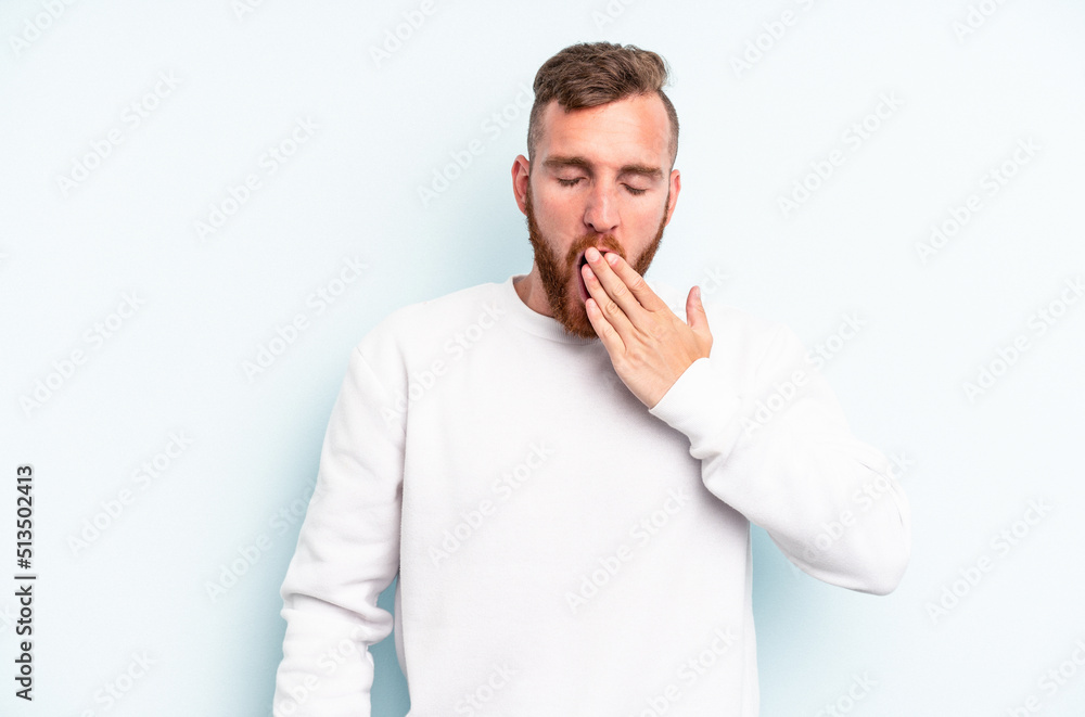 Young caucasian man isolated on blue background yawning showing a tired gesture covering mouth with hand. Young caucasian man isolated on blue background yawning showing a tired gesture covering mouth with hand.