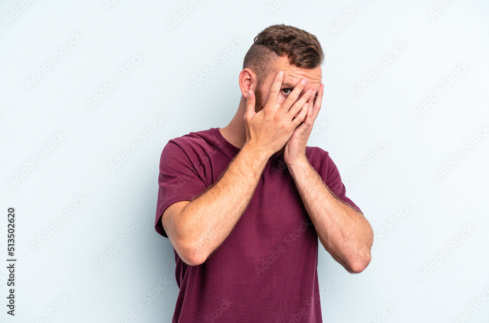 Young caucasian man isolated on blue background blink at the camera through fingers, embarrassed covering face. Young caucasian man isolated on blue background blink at the camera through fingers, embarrassed covering face.
