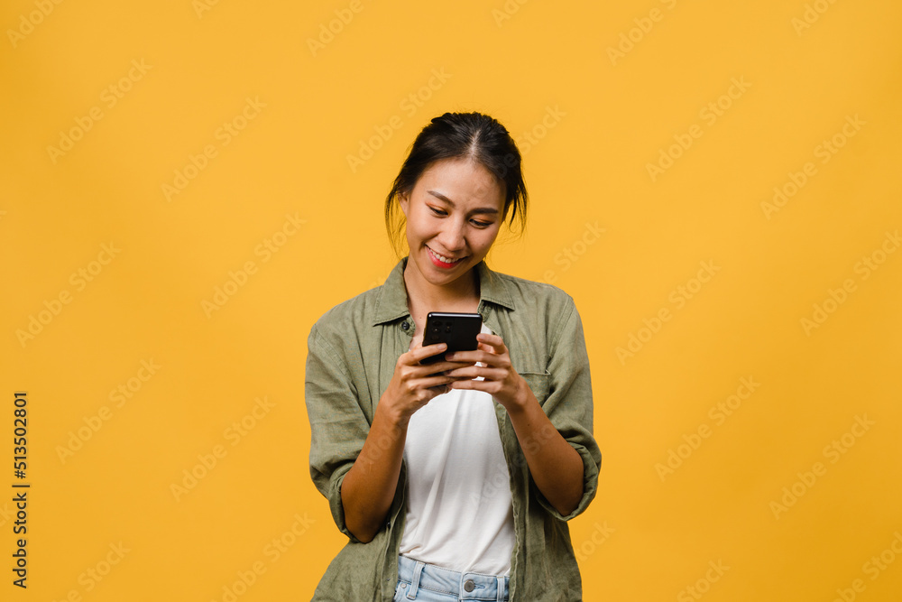 Young Asia lady using phone with positive expression, smiles broadly, dressed in casual clothing feeling happiness and stand isolated on yellow background. Happy adorable glad woman rejoices success. Young Asia lady using phone with positive expression, smiles broadly, dressed in casual clothing feeling happiness and stand isolated on yellow background. Happy adorable glad woman rejoices success.