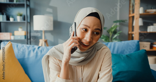 Attractive cheerful young Asian muslim beauty woman in hijab with casual use phone call with friend and family in cozy living room at home. Girl Islam social distance quarantine lifestyle concept.
