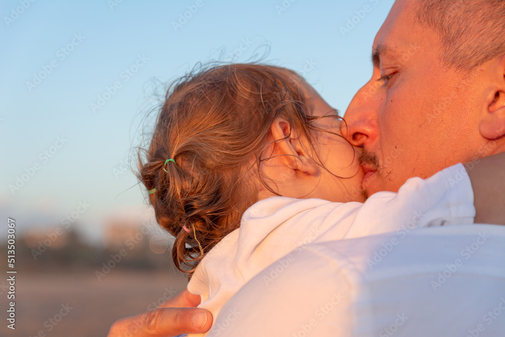 A young dad with a little daughter walks along the sea beach in the sunset.