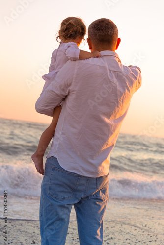A young dad with a little daughter walks along the sea beach in the sunset.