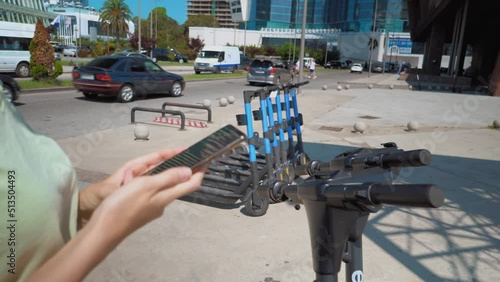 Close-up of woman's hands holding phone near modern city electric scooter, about to unlock it. Modern urban transport. uses eco-transport in city center. view from back.