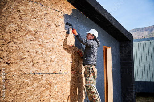 Male painter using paint roller, doing exterior paint work in a black color. Man worker building wooden frame house. Carpentry and construction concept. © anatoliy_gleb