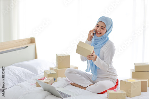Beautiful and young asian woman in muslim sleepwear with attractive look, sitting on bed with computer and online package box delivery. Startup small business SME freelance girl work with mobile phone