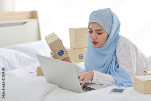 Beautiful and young asian woman in muslim sleepwear with attractive look, lies on bed with computer and online package box delivery. Startup small business SME freelance girl working with mobile phone © feeling lucky