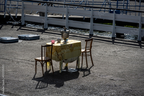 a table with a tablecloth and a samovar on a platform at the pier of ships on a summer day