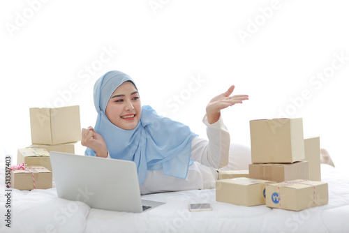 Beautiful and young asian woman in muslim sleepwear showing something, lies on bed with computer and online package box delivery. Startup small business SME freelance girl working with mobile phone © feeling lucky