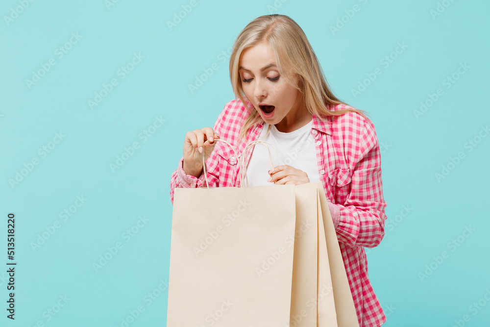 Fototapeta premium Young amazed woman she 30s in pink shirt white t-shirt hold look at package bags with purchases after shopping isolated on plain pastel light blue background studio portrait. People lifestyle concept.