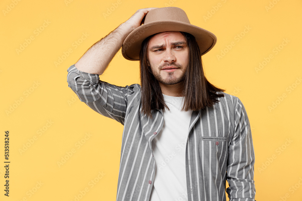 Young sad confused man he 20s wears striped grey shirt white t-shirt hat look aside on workspace hold scratch head isolated on plain yellow color background studio portrait. People lifestyle concept. Young sad confused man he 20s wears striped grey shirt white t-shirt hat look aside on workspace hold scratch head isolated on plain yellow color background studio portrait. People lifestyle concept.