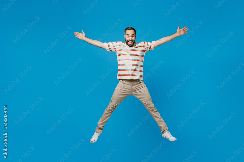 Full body young overjoyed excited surprised man 20s in orange striped t-shirt looking camera jump high with outstretched hands legs isolated on plain blue background studio. People lifestyle concept.