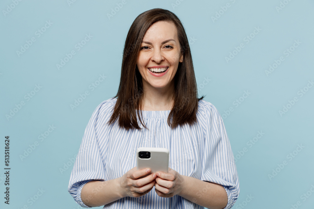 Young happy smiling fun woman she 20s wears casual blouse hold in hand use mobile cell phone browsing internet isolated on pastel plain light blue background studio portrait. People lifestyle concept. Young happy smiling fun woman she 20s wears casual blouse hold in hand use mobile cell phone browsing internet isolated on pastel plain light blue background studio portrait. People lifestyle concept.