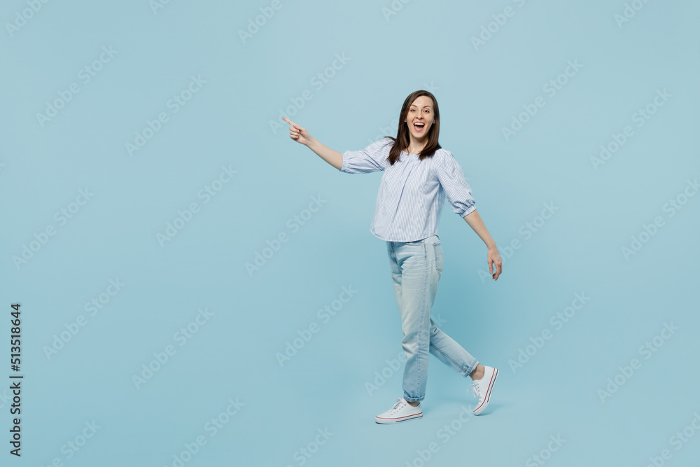 Full body side view young happy smiling woman she 20s in casual blouse walking going point index finger aside isolated on pastel plain light blue background studio portrait. People lifestyle concept.