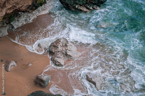 Aerial close-up view of rocky shore with crashing sea waves and sand. Atlantic ocean in the coast of Portugal