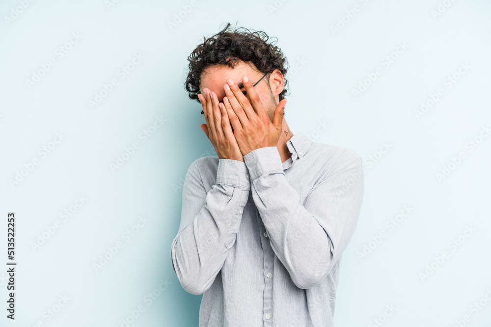 Young caucasian man isolated on blue background blink at the camera through fingers, embarrassed covering face. Young caucasian man isolated on blue background blink at the camera through fingers, embarrassed covering face.