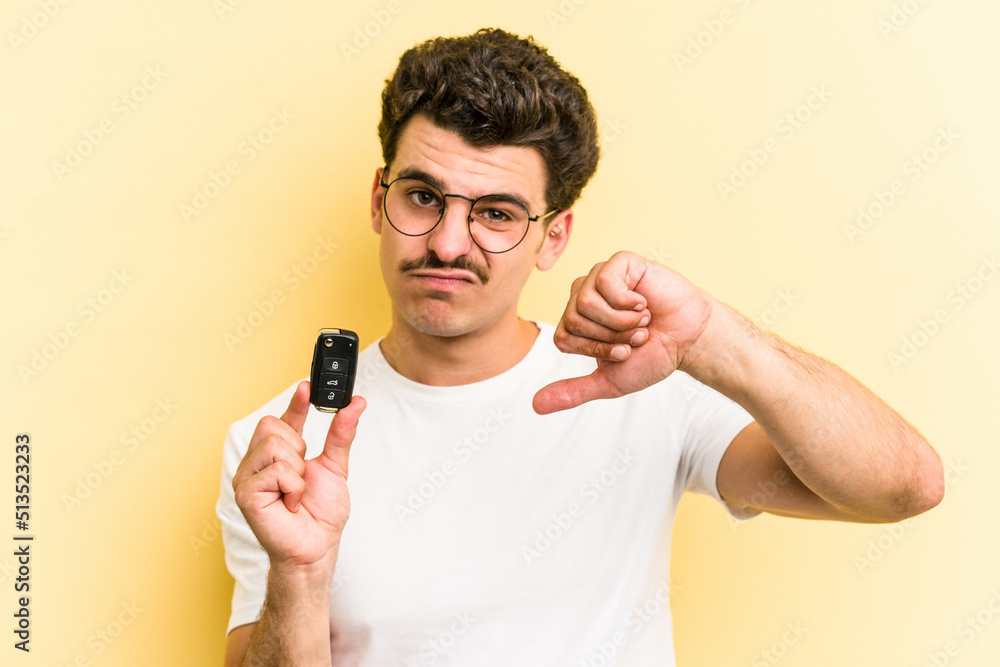 Young caucasian man holding car keys isolated on yellow background showing a dislike gesture, thumbs down. Disagreement concept.
