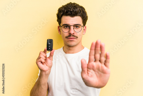 Young caucasian man holding car keys isolated on yellow background standing with outstretched hand showing stop sign, preventing you. © Asier