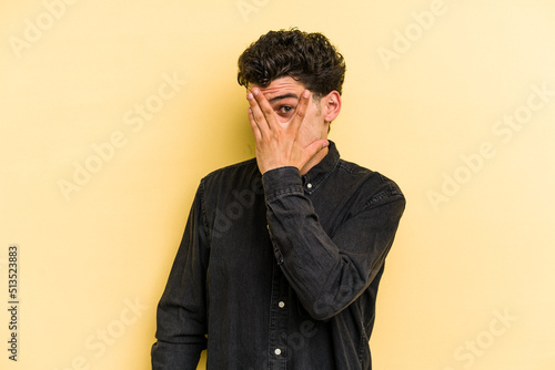 Young caucasian man isolated on yellow background blink at the camera through fingers, embarrassed covering face. © Asier