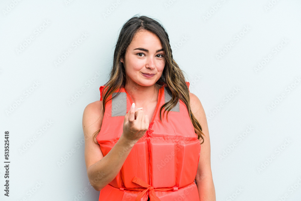 Young caucasian woman wearing life jacket isolated on blue background pointing with finger at you as if inviting come closer. Young caucasian woman wearing life jacket isolated on blue background pointing with finger at you as if inviting come closer.