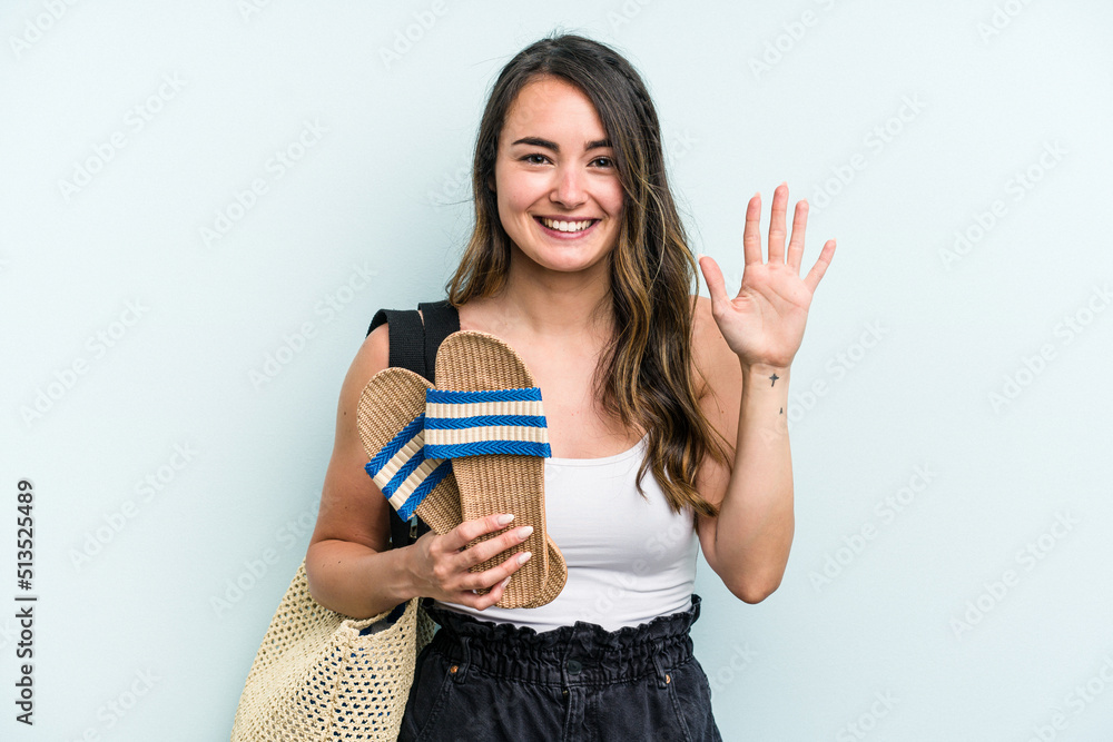 Young caucasian woman holding sandals isolated on blue background smiling cheerful showing number five with fingers. Young caucasian woman holding sandals isolated on blue background smiling cheerful showing number five with fingers.
