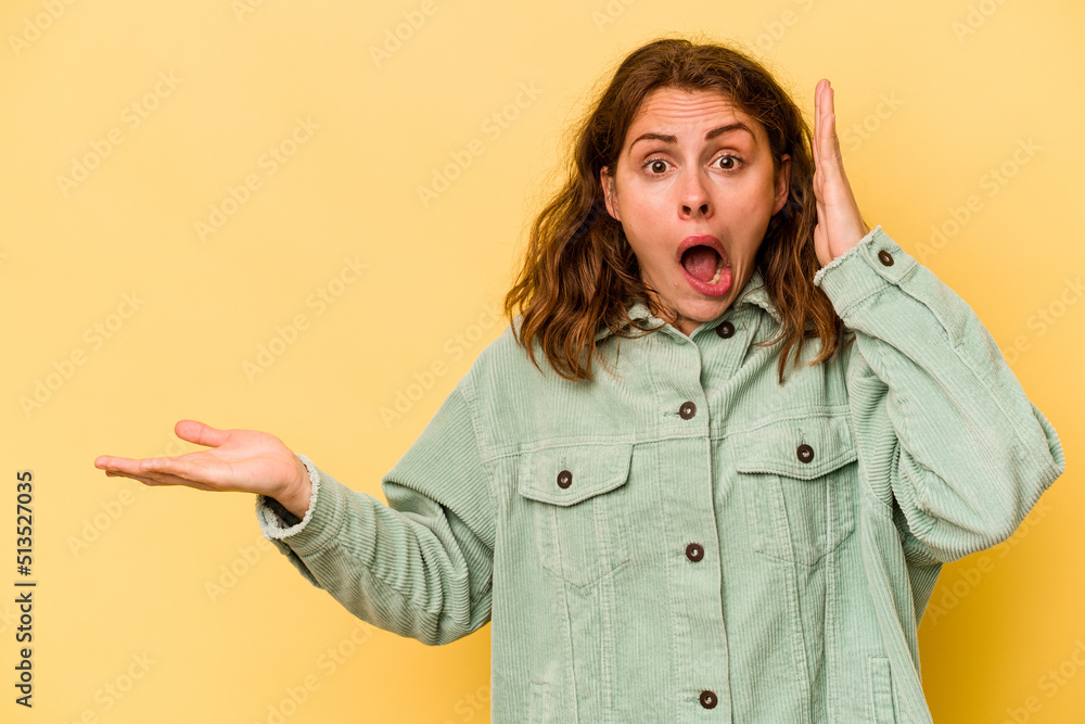 Young caucasian woman isolated on yellow background holds copy space on a palm, keep hand over cheek. Amazed and delighted. Young caucasian woman isolated on yellow background holds copy space on a palm, keep hand over cheek. Amazed and delighted.