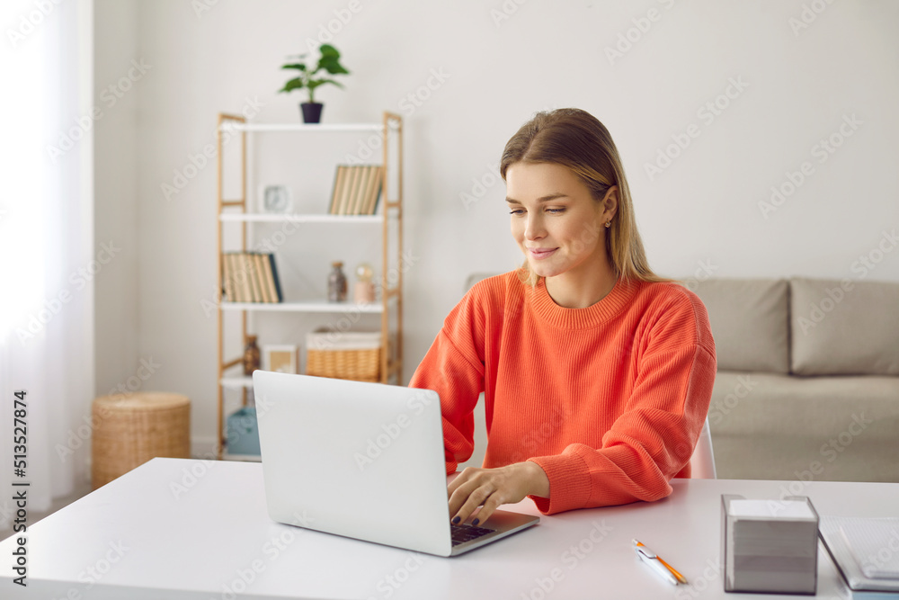 Ambitious young woman working remotely or distance learning at home using laptop. Smiling caucasian girl typing on laptop while sitting at desk in home office. Freelance or studying concept.