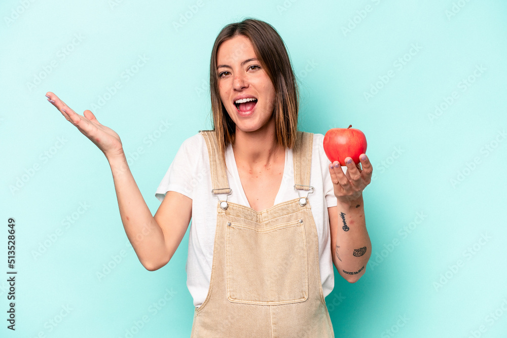 Young caucasian pregnant woman holding an apple isolated on blue background receiving a pleasant surprise, excited and raising hands.