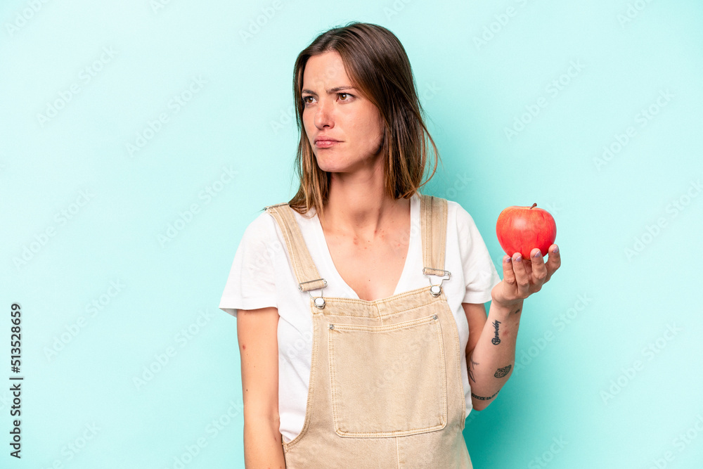 Young caucasian pregnant woman holding an apple isolated on blue background confused, feels doubtful and unsure.