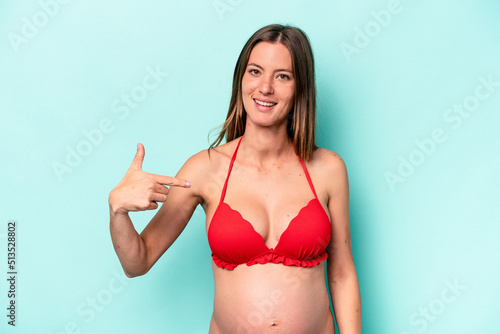 Young caucasian pregnant woman wearing bikini isolated on blue background person pointing by hand to a shirt copy space, proud and confident © Asier