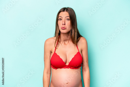 Young caucasian pregnant woman wearing bikini isolated on blue background shrugs shoulders and open eyes confused. © Asier
