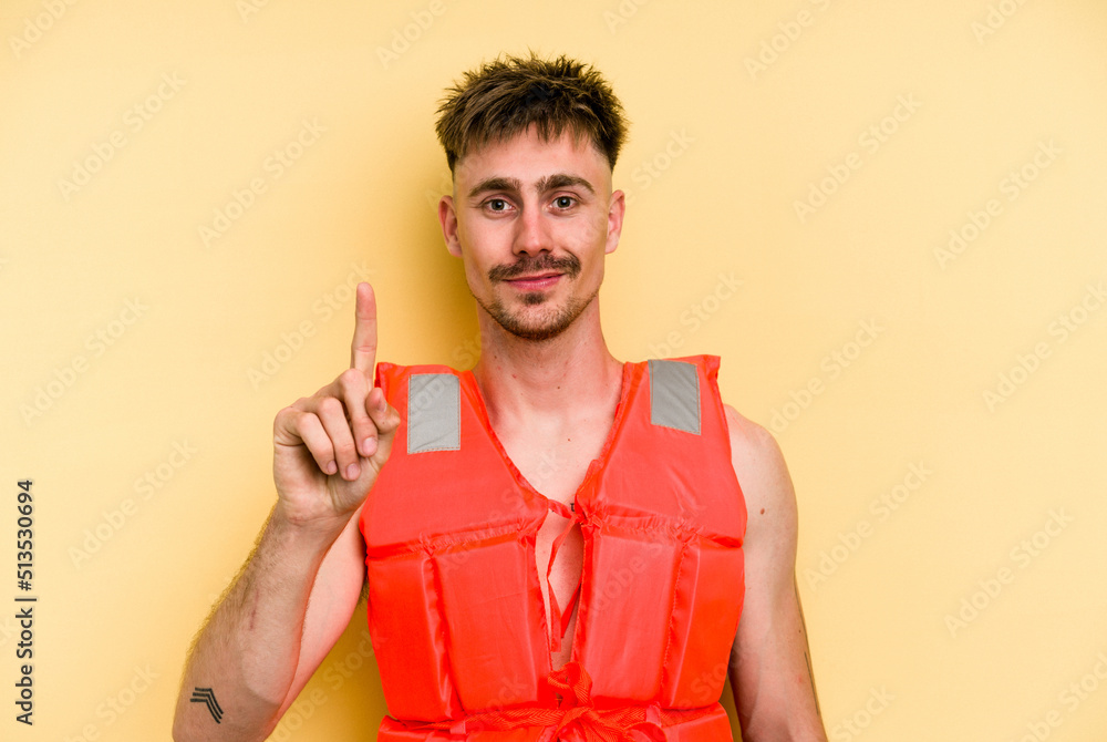Young caucasian man wearing a life jacket isolated on yellow background showing number one with finger. Young caucasian man wearing a life jacket isolated on yellow background showing number one with finger.