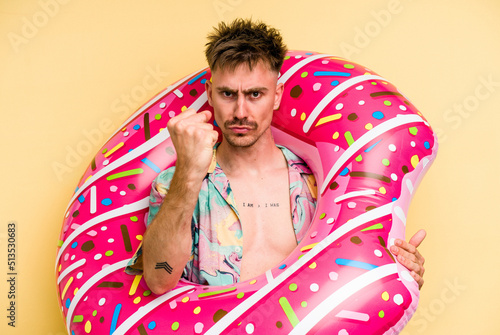 Young caucasian man holding an inflatable donut isolated on yellow background showing fist to camera, aggressive facial expression. © Asier