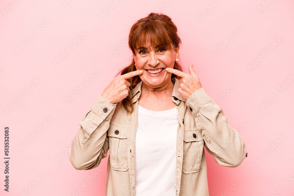Middle age caucasian woman isolated on pink background smiles, pointing fingers at mouth.