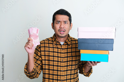 Adult Asian man showing worried expression when holding paper money and pile of stuff boxes photo