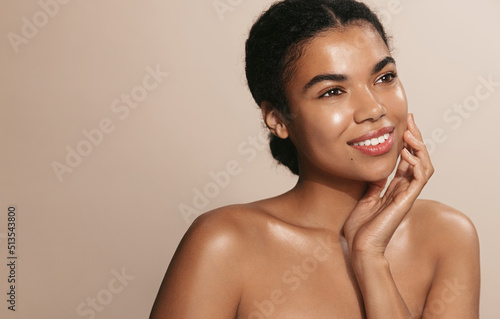 Beauty female model with nourished, glowing skin, smiling, gently touching her moisturized face, brown background