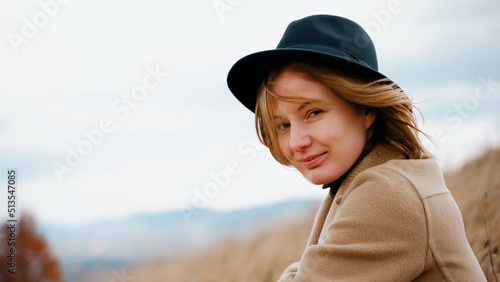 Portrait of beautiful smiling girl looking into camera blinks eyes. Woman in hat on head in open air in windy weather on background of mountains and sky. Lightness at soul, spiritual calm and freedom