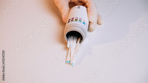 Close-up of hand in disposable medical glove pour out test strips from plastic jar on white background. Strips for urine diagnosis. Test for glucose and acetone. Strips for self-diagnosis. Laboratory