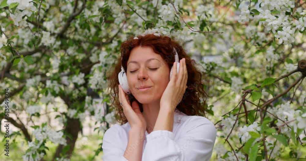 Red-haired woman in headphones in a flowering garden listening to music while dancing Red-haired woman in headphones in a flowering garden listening to music while dancing