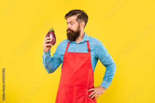 Fotografia Serious man in red apron looking at eggplant yellow background, grocer