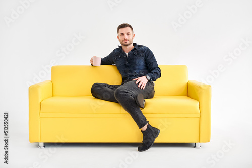 Relaxed man with coffee mug in hands sitting on comfort bright sofa looking to camera